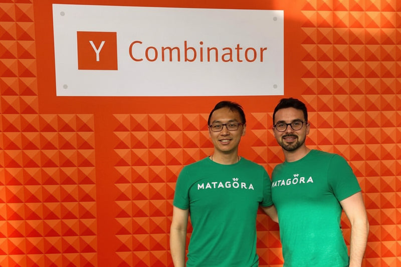 Two people wearing green 'MATAGORA' shirts standing in front of a 'Y Combinator' sign on an orange wall.